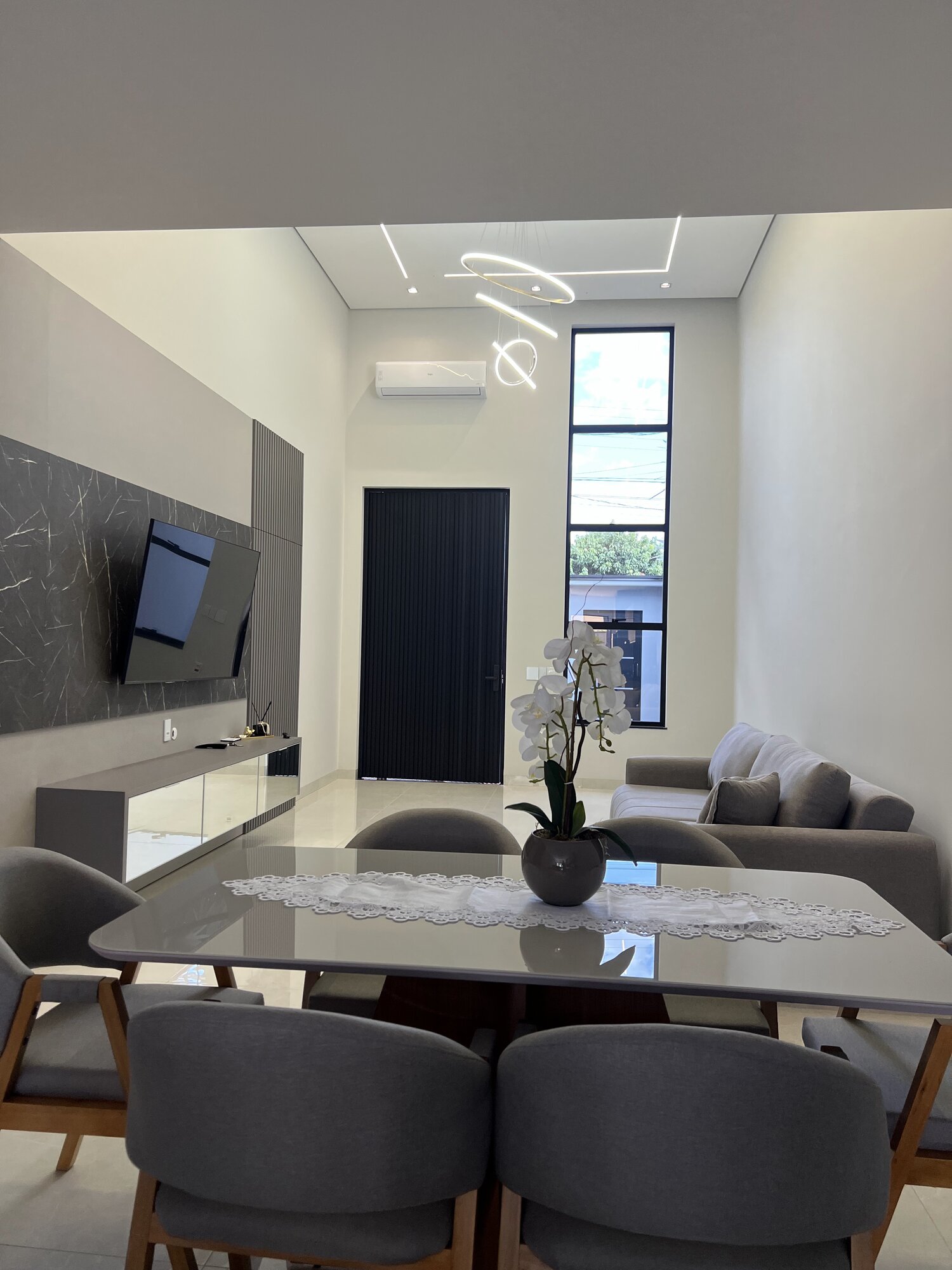 Sala de jantar integrada ao living com mesa para seis lugares, pendente em anéis luminosos e pé-direito duplo.