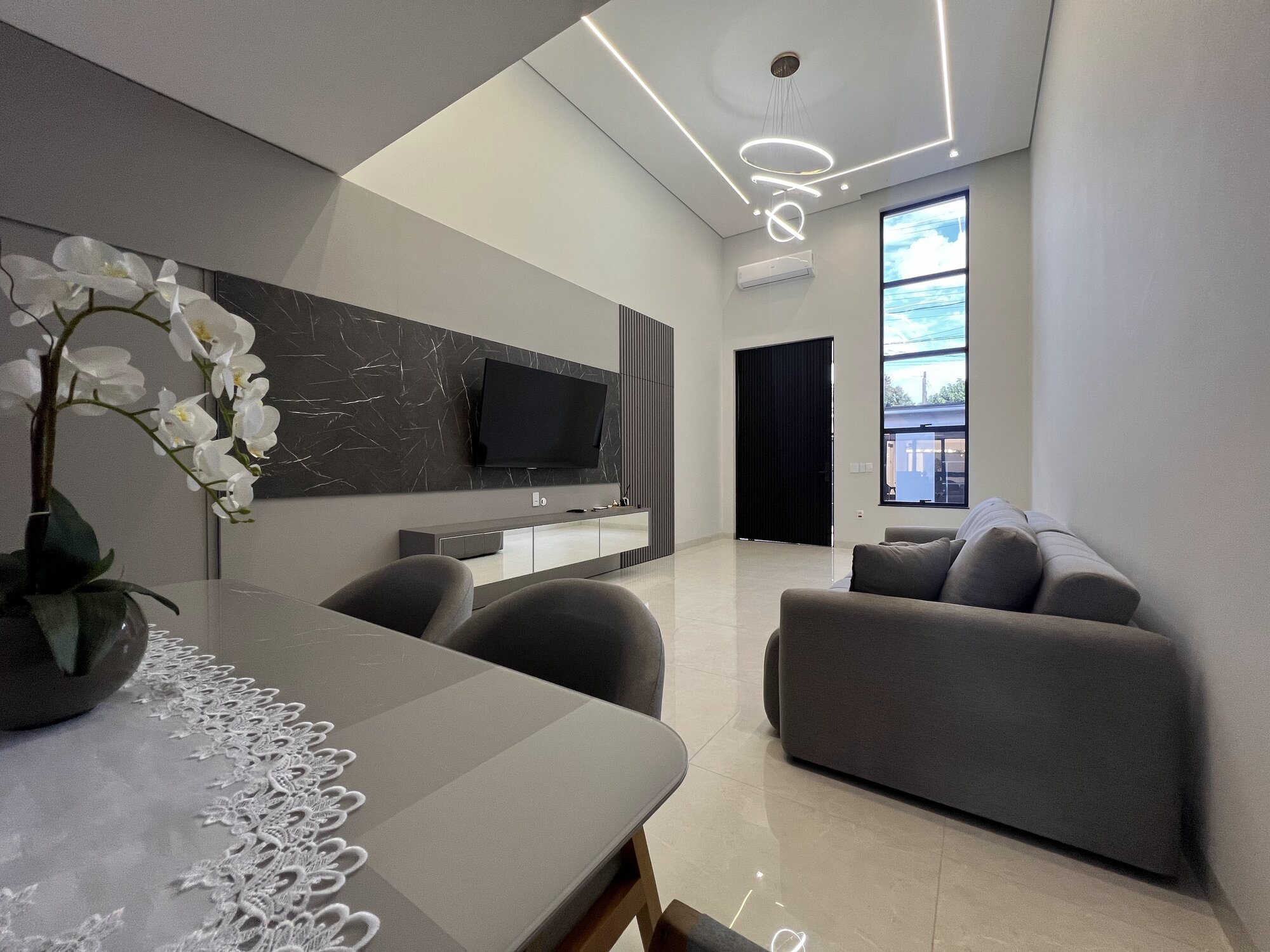 Living com painel de TV em mármore escuro, sofá cinza, rack espelhado e iluminação linear no forro.