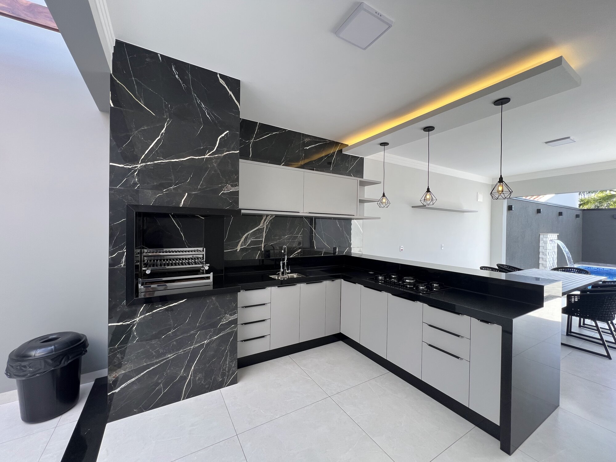 Espaço gourmet com churrasqueira em mármore preto, bancada em granito, pendentes industriais e vista para a piscina.